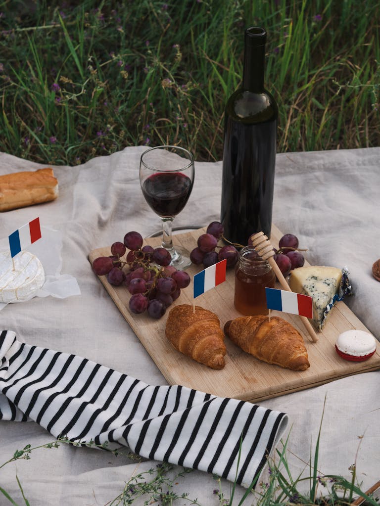 A delicious French picnic setup with wine, cheese, grapes, and pastries on a blanket.