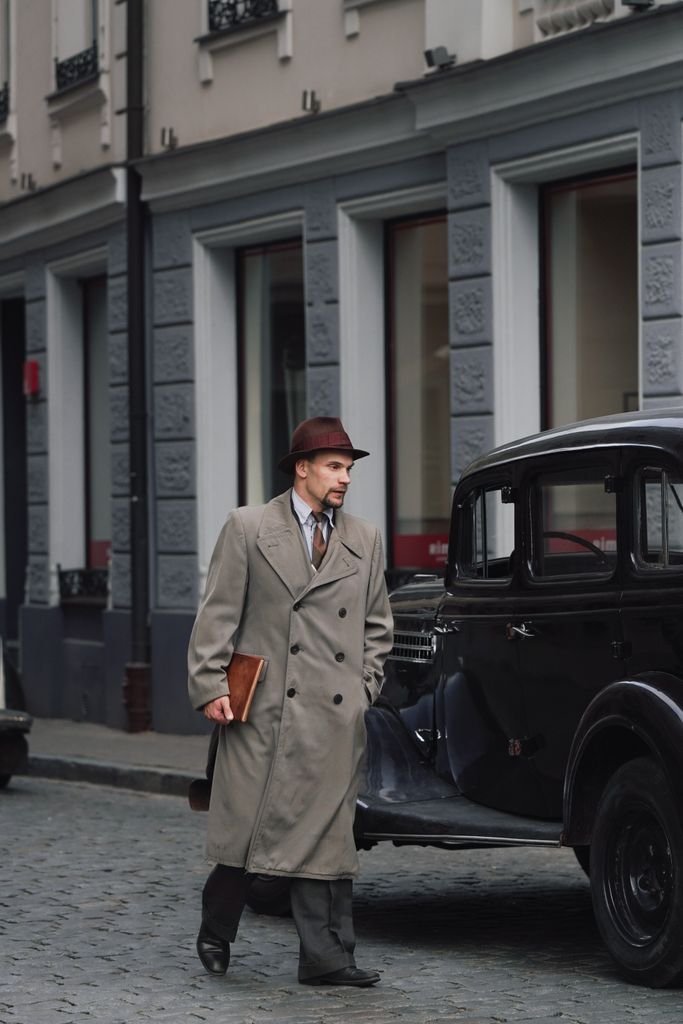 A man in a vintage coat and hat walking past a classic car on a historic Riga street.
