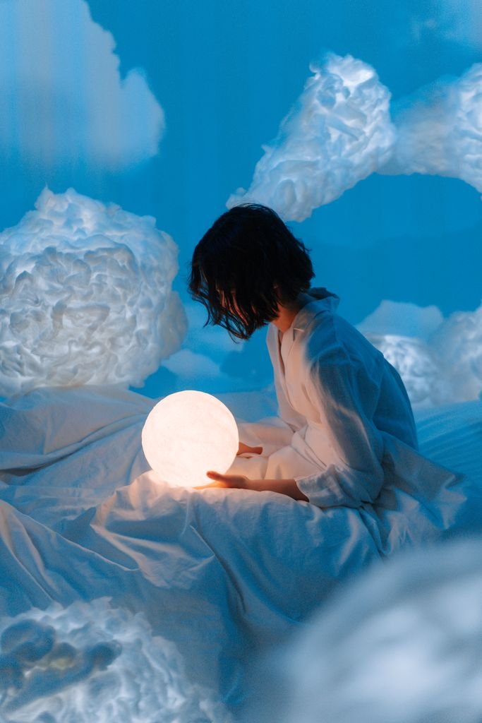 A serene scene of a woman holding an illuminated moon-shaped lamp in a dreamy, cloud-like setting.