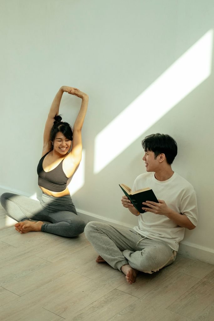 Full body of positive Asian couple in activewear sitting on floor with crossed legs while smiling and looking at each other