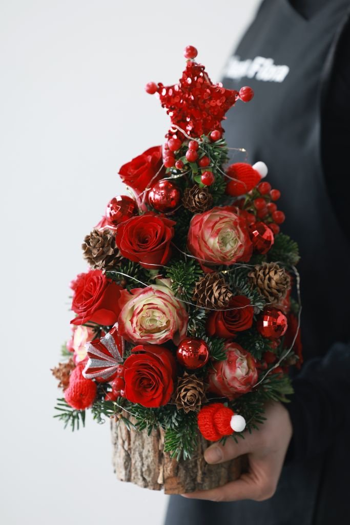 Hand holding a Christmas-themed floral arrangement with red roses and pine cones.